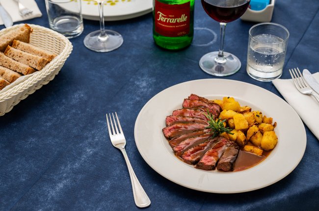 Piatto di carne e patate con bicchieri, posate e pane.