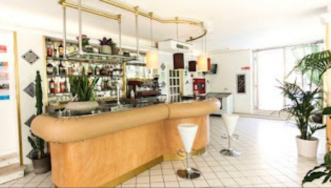 Angolo bar con bancone curvo, sgabelli bianchi e decorazioni vegetali. Bottiglie sugli scaffali.