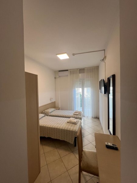 Stanza d'hotel con due letti singoli, finestra con tende bianche, TV a muro e scrivania.