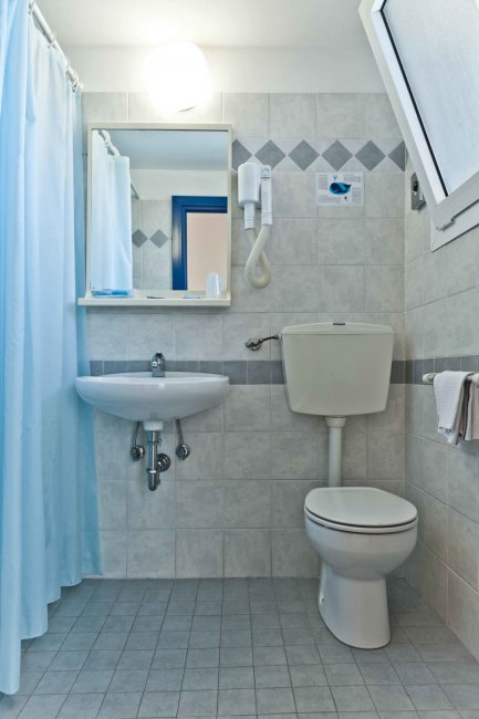 Un bagno piastrellato con lavabo, wc, specchio e asciugacapelli. Doccia con tenda azzurra visibile.