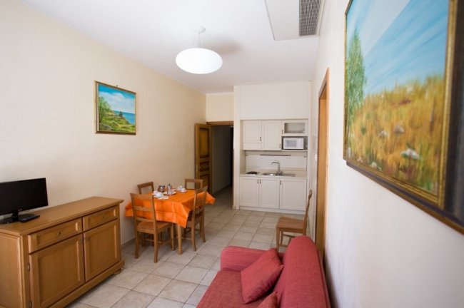 Soggiorno con divano rosso, tavolo arancione e piccola cucina.