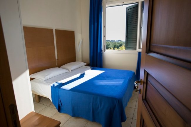 Letto matrimoniale con copriletto blu, finestra aperta.