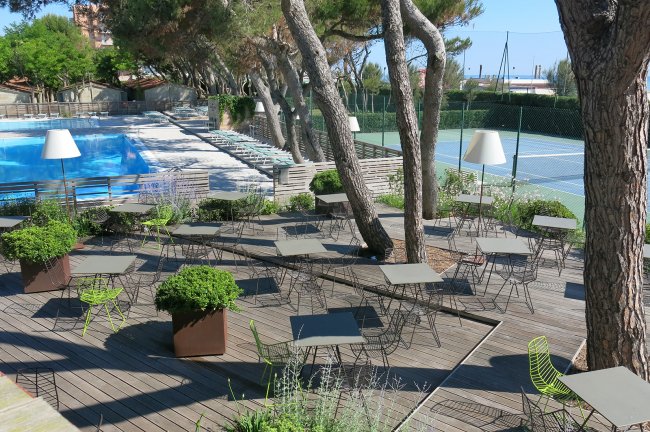 Terrazza con sedie e tavolini vicino a piscina e campo da tennis.