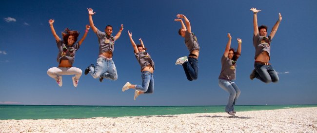 Sei persone saltano sulla spiaggia sotto un cielo blu.