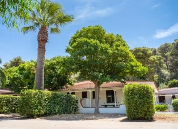 Casetta bianca circondata da alberi e cespugli, con cielo sereno.