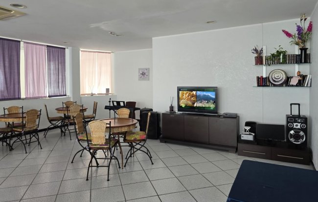 Sala con tavoli rotondi e sedie di vimini, TV su mobile scuro e scaffale con decorazioni.