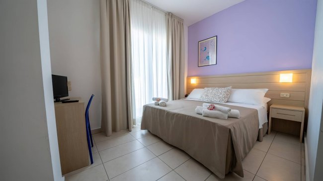 Camera da letto semplice con letto matrimoniale, parete lilla e finestra con tenda bianca.